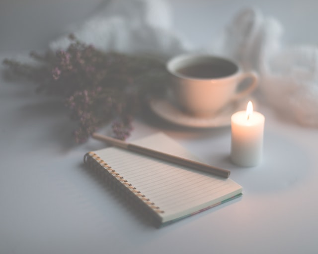 A cozy scene with a journal on a table next to a lit white candle, cup of coffee, and purple flowers.