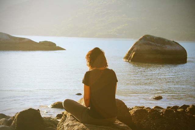 Lady sitting at edge of calm waters looking at the view with rocks dispersed within the calm waters.