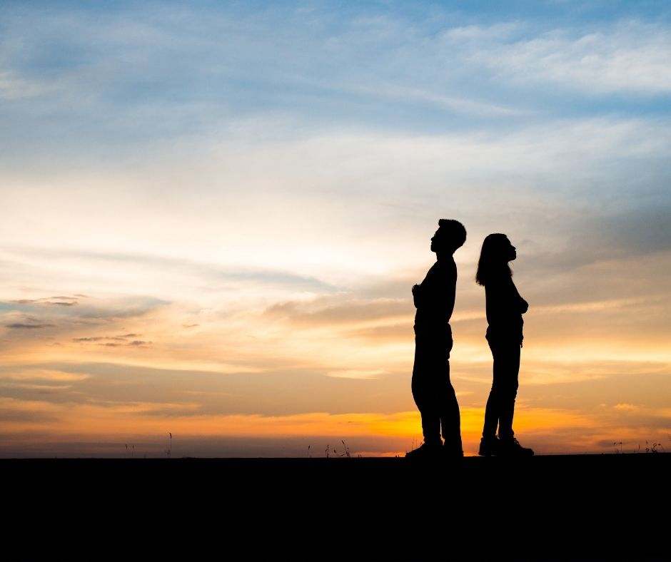 Couple with their backs to eachother in front of a sunset.
