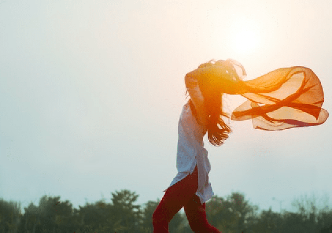 Woman feeling free with a red, sheer scarf flowing behind her as she grasps it by her shoulders, under the sunset.