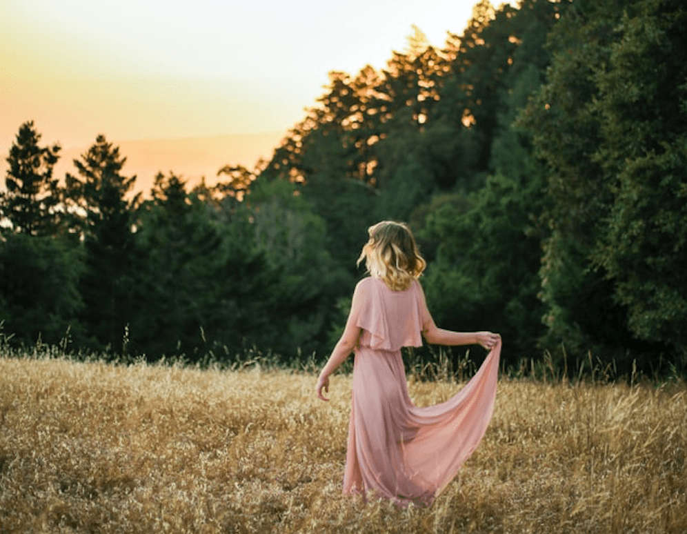 Lady in a pink dress walking through a field with pine trees in the distance at sunrise