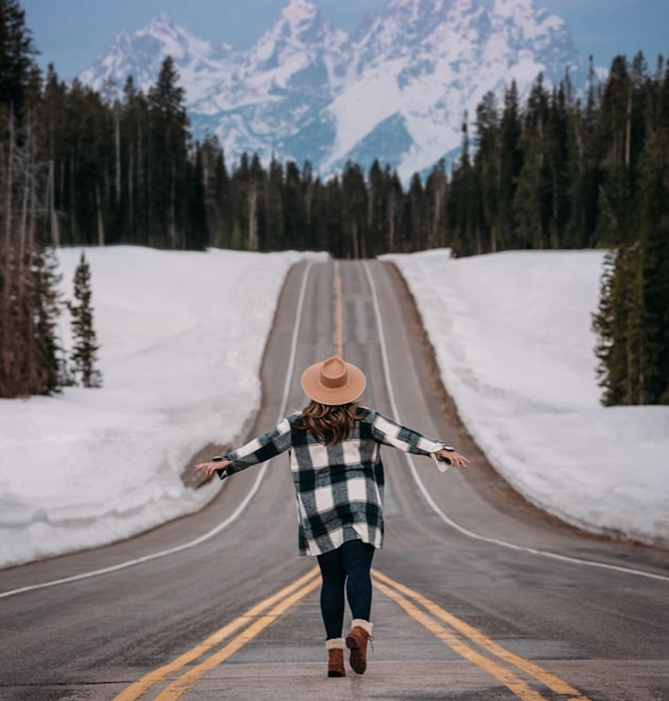 Woman walking down a road with no traffic and mountains that look like the Himalayas ahead. It is so gorgeous! Pine trees all around.