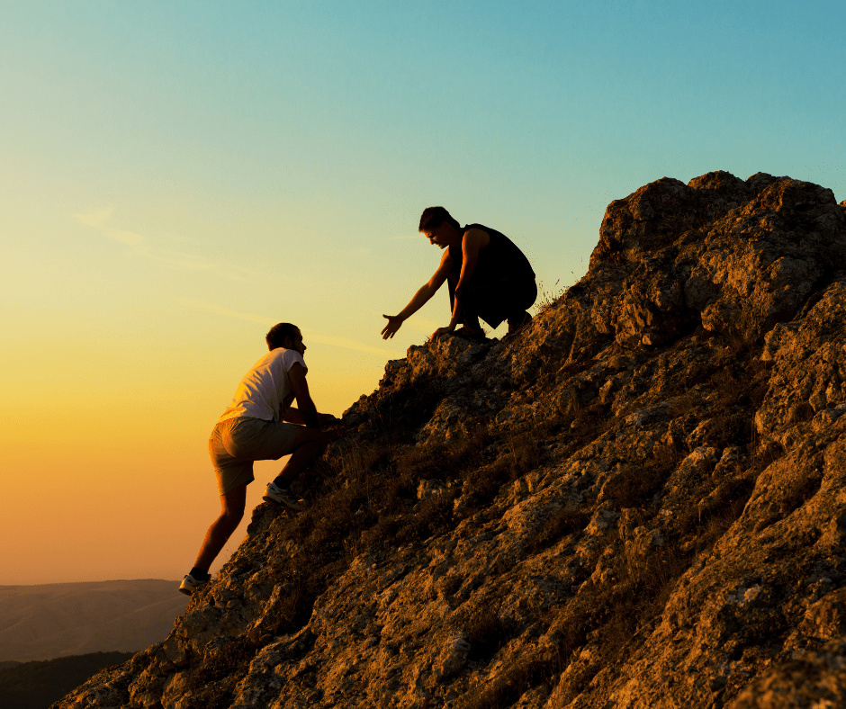 man reaching out his hand to another man on the side of a mountain